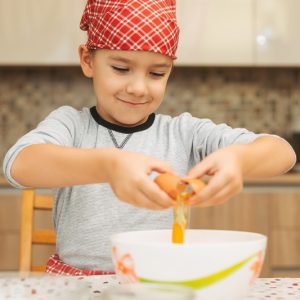 Smiling 7 year old boy helping in the kitchen, breaking eggs into large bowl.